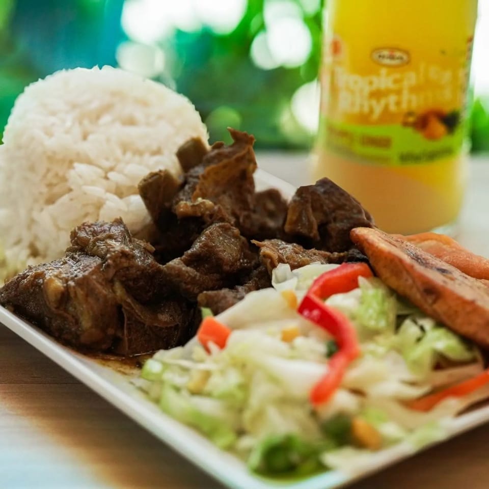 Grilled meat served with white rice, roasted plantains, fresh salad, and a golden beverage.