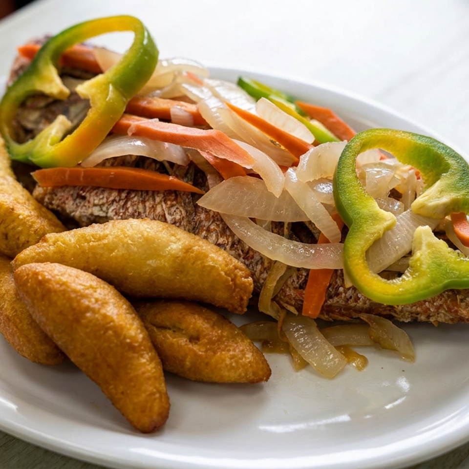 Whole fried fish topped with sautéed onions, carrots, and green peppers, served with golden fried plantains on a white plate.