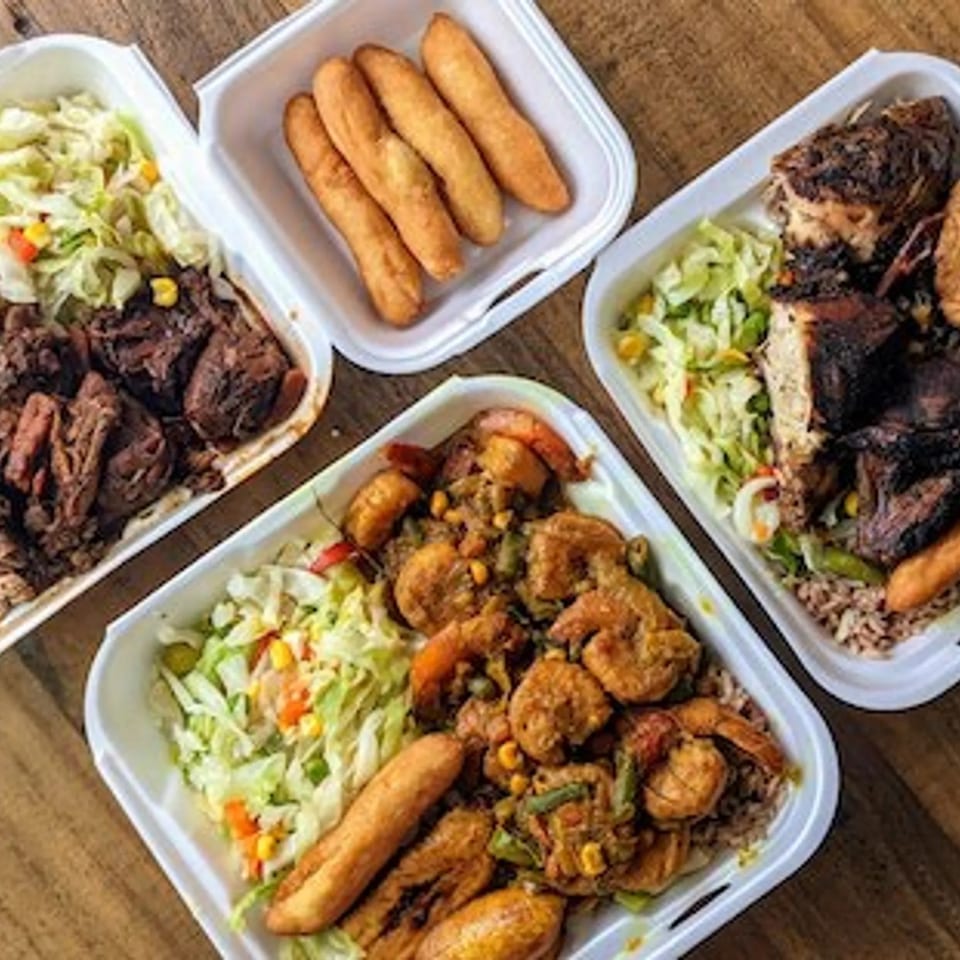 Assorted takeout meals with grilled meat, fried plantains, rice, salad, and golden croquettes arranged on a wooden table.